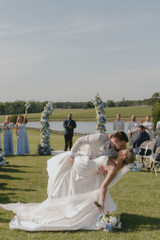 Newlywed couple shares a dramatic kiss dip during an outdoor lakeside ceremony, framed by floral pillars filled with blue, white, and green blooms, while guests and wedding party look on.