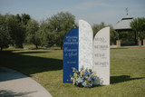 Modern blue and white wedding welcome display featuring custom signage and a floral arrangement of blue, white, and green blooms set along a grassy outdoor venue entrance.