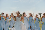 Bride poses with her bridesmaids in light blue dresses, each carrying blue, white, and green bouquets while holding white parasols during an outdoor wedding celebration.