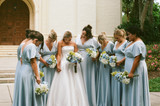 Bride surrounded by seven bridesmaids in light blue dresses outside a church entrance, all holding blue, white, and green hydrangea bouquets and looking down in a quiet, candid moment before the ceremony.