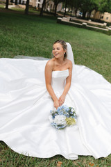 Bride seated on the flowing skirt of her wedding gown outdoors, smiling while holding a bouquet of blue, white, and green hydrangeas and coordinating florals.
