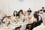 Guests laugh and talk amongst one another sitting down at rectangle table with light pink and ivory centerpieces in gold scalloped vases lining the table down the center.