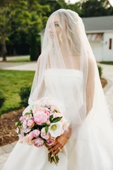 Bride holds a light pink and white bridal bouquet with a veil covering her face as she soft smiles and looks into the distance.