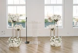 A mix of white floral arrangements in different sizes and designs arranged on stands and on the floor for a stylish wedding ceremony backdrop.