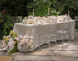 Outdoor reception table with a soft ivory linen, clear Chiavari chairs, and lush floral runners featuring blush anthuriums, white orchids, pale pink roses, and soft green hydrangeas. A swan planter overflowing with blooms sits beside the table.