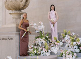 Two bridesmaids in mocha and blush satin dresses stand among abundant floral installations of blush anthuriums, white orchids, pale pink roses, and soft green hydrangeas while holding white calla lily bouquets.