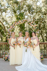 Bride posing with bridesmaids in pale yellow dresses beneath a circular floral arch decorated with peach and blush flowers.