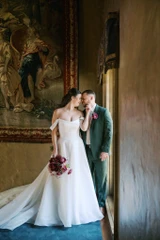 A bride holding a bouquet made up of different shades of purple florals and groom wearing a emerald green suit pose for a photo indoors with a tapestry in the background.