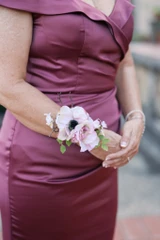 A mother of the bride wears a satin purple dress with a wrist corsage featuring a lilac anemone.