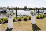 Two light blue and white floral meadows are placed on top of two white pillars at a wedding ceremony overlooking a boat harbor with water and boats in the background.