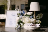 A light blue and white floral vaseless centerpiece sits on top of a bar next to a framed bar menu, small table side lamp, and a bowl shaped like a clam shell that holds matchboxes for the guests to take as a favor.