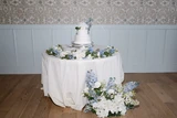 A cake table at a wedding reception with a light blue and white floral garland encircles the cake and a complimentary floral meadow sits at the base of the table.