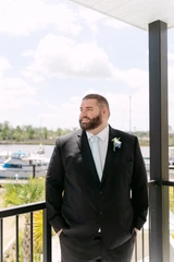A groom poses and looks off into the distance wearing a black suit with a white rose boutonniere pinned to his jacket with a harbor and boats as the backdrop of the photo.