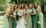 Bride in a white lace wedding gown poses outdoors with bridesmaids in assorted green dresses, all holding white floral bouquets in a wooded garden setting.