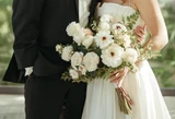 Bride in a white wedding gown holds a bouquet of white flowers and greenery while standing beside the groom in a black tuxedo.