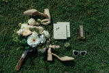 Wedding flat lay on green grass featuring a bridal bouquet of white flowers, nude heels, perfume, sunglasses, and an invitation suite.