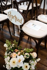 Reserved seating sign displayed on a wooden ceremony chair beside a floral arrangement of white and blush flowers inside a wedding venue.