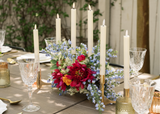Colorful Kinsley Collection silk floral centerpiece featuring vibrant pink peonies, blue delphinium, white wildflowers, and orange accents arranged among ivory taper candles. Crystal glassware, gold candleholders, and elegant place settings complete the cheerful garden-inspired wedding tablescape.