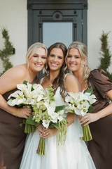 Bride holding a white and green floral arrangement poses with her bridesmaids wearing brown dresses and holding a classic and modern all white calla lily and anthurium bouquets.