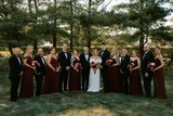 A bride and groom pose with their wedding party, bridesmaids are wearing burgundy and groomsmen are wearing black tuxes, all while holding deep red, mauve, and ivory bouquets.