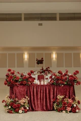 A close-up of a sweetheart table at a wedding reception with red crushed velvet linens and deep red, mauve, and ivory floral meadows placed intentionally on and at the base of the table.
