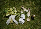 A flat lay on grass of a classic green and white bridal bouquet style with white bridal shoes, wedding rings, a perfume bottle, and his and her vow books.