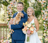 Bride and groom posing with their dog in front of a peach and blush floral arch while holding a romantic wedding bouquet.