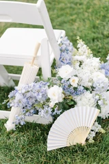 A light blue and white floral meadow sits at the base of a chair at a wedding ceremony with a parasol and hand fan placed next to it for a styled image.