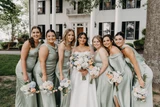 A bride and her bridesmaids wearing silk sage dresses pose for a photo while holding multi-color pastel floral bouquets.