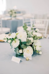 A white and greenery small vaseless floral arrangement is placed in the center of a round table with a light blue tablecloth next to a "reserve" sign.
