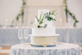 A two tier classic white cake is decorated with white florals and greenery and sits on top of a muted gold cake stand placed on top of a round table with a light blue and lace tablecloth.