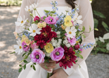 lose-up of the Kinsley Collection bridal bouquet featuring vibrant silk wildflowers, including white cosmos, deep burgundy peonies, pink ranunculus, yellow poppies, blue delphinium, and delicate white filler flowers. The colorful hand-tied bouquet is held by a bride in a white wedding gown.