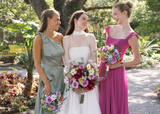 Bride in a white gown stands between two bridesmaids in sage green and fuchsia dresses, each holding colorful Kinsley Collection silk bouquets featuring pink roses, blue delphinium, white daisies, yellow blooms, and vibrant wildflowers. The bridal party smiles together in a lush garden setting.