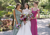 Bride in a white gown stands between two bridesmaids in sage green and fuchsia dresses, each holding colorful Kinsley Collection silk bouquets featuring pink roses, blue delphinium, white daisies, yellow blooms, and vibrant wildflowers. The bridal party smiles together in a lush garden setting.