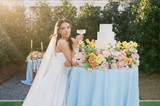 Bride in a modern white gown and lace-edged veil poses beside a three-tier white cake adorned with lush florals in shades of blush, peach, yellow, and soft green.