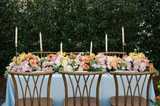 Romantic reception table styled with pale blue linens, wooden chairs, and abundant floral runners featuring blush and peach roses, yellow tulips, pale pink ranunculus, and soft greenery.