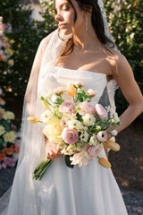 Bride in a modern white gown and lace-edged veil holds a garden-inspired bouquet of pale pink ranunculus, blush and cream roses, yellow tulips, and delicate white blooms.