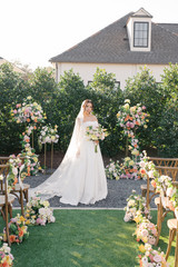 Bride in a modern white gown and flowing lace-edged veil stands between two floral ceremony arches overflowing with blush, peach, yellow, and ivory blooms, holding a coordinating garden-inspired bouquet.