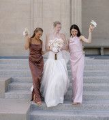 Bride in a modern white gown and veil walks down stone steps between two bridesmaids in mocha and blush satin dresses. The bride carries a bouquet of blush anthuriums, white orchids, and soft ivory blooms, while the bridesmaids hold petite white floral posies.