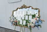 Two multi-colored pastel floral swags are adhered to a seating chart made out of an gold ornate trim mirror.