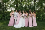 Bride stands with her bridal party wearing light pink bridesmaid dresses and one man in a light tan suit. The bride holds light pink and white bridal bouquet and the bridesmaids hold a petite bouquet made up of lavender roses.