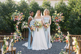 A bride and her bridesmaids in blue satin dresses holding floral bouquets and surrounded by florals