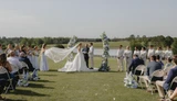 Bride poses with groom in light blue dresses, each carrying blue, white, and green bouquets while standing in front of a split asymmetrical ceremony installation for an outdoor wedding celebration.