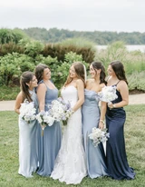 A bride and her bridesmaids wearing different shades of blue look at each other laughing as they pose for a photo holding light blue and white bouquets.