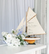 A light blue and white floral vaseless centerpiece sits on top of a bar next to a miniature sail ship with the cocktail menu written in script on the sail.