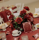 Elegant wedding reception table with gold chiavari chairs and a dramatic centerpiece of red roses, burgundy calla lilies, and candles on a red linen tablecloth.