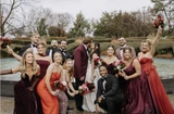 A bride and groom share a kiss while their wedding party wearing shades of pink, red, and maroon cheer them on at an outdoor wedding ceremony.
