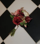 A deep red, mauve, and ivory bridesmaid bouquet lays flat on a black and white checkered floor.