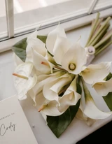 Modern Callas Lily bridal bouquet is laid flat on a windowsill next to a vow booklet.