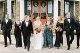 A wedding party with the grooms in black tuxes and girls in emerald green bridesmaid dresses walk forward holding red and pink bouquets